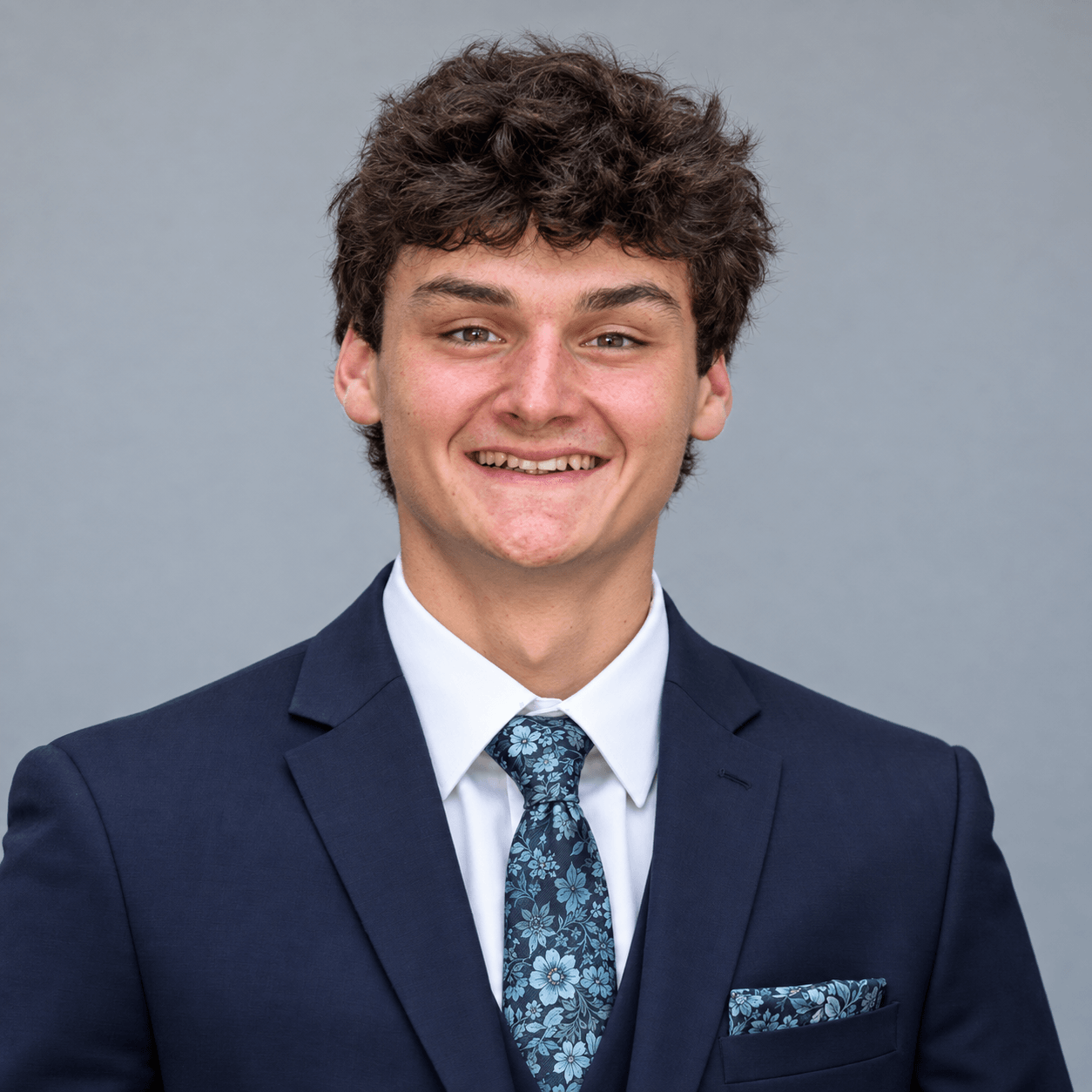 Ross Champlin smiling in a navy suit and floral tie against a gray studio backdrop